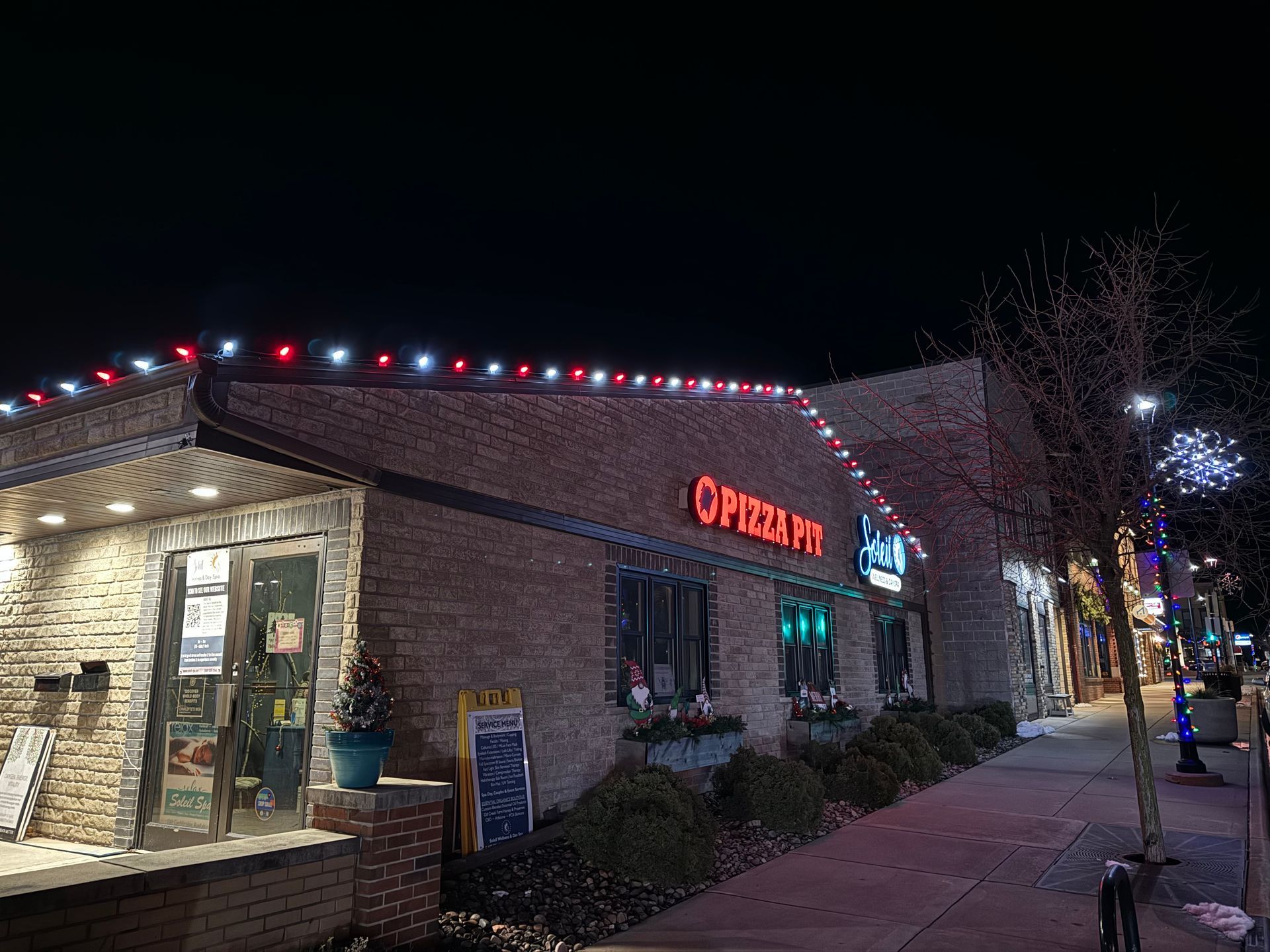 Nighttime sidewalk view of a lit brick restaurant with red and white roof lights and a glowing sign.