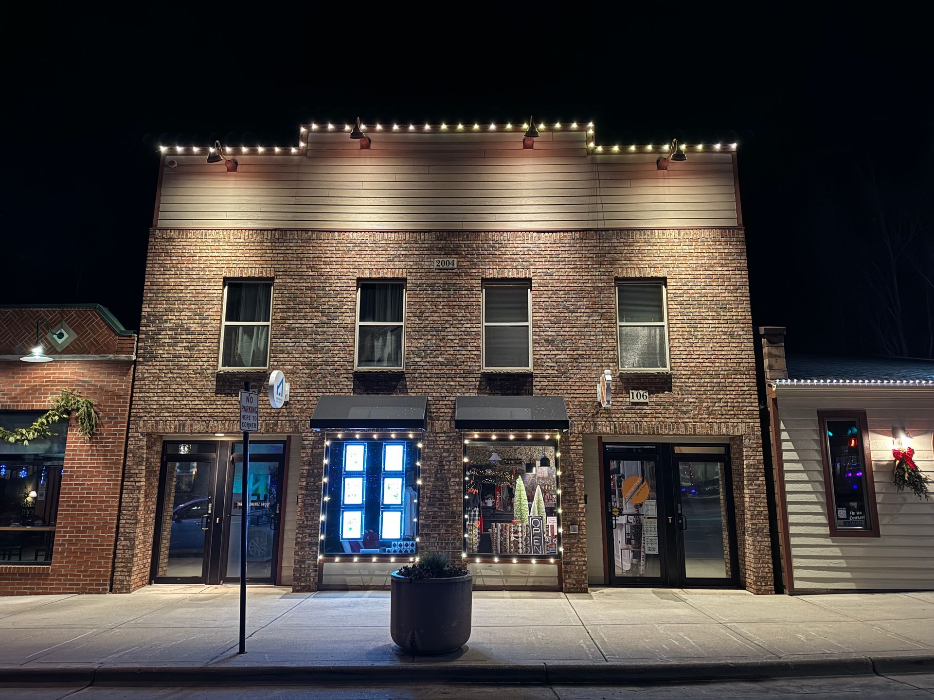 Brick storefront lit at night with blue-lit central doors, holiday decorations, and snow on the sidewalk.