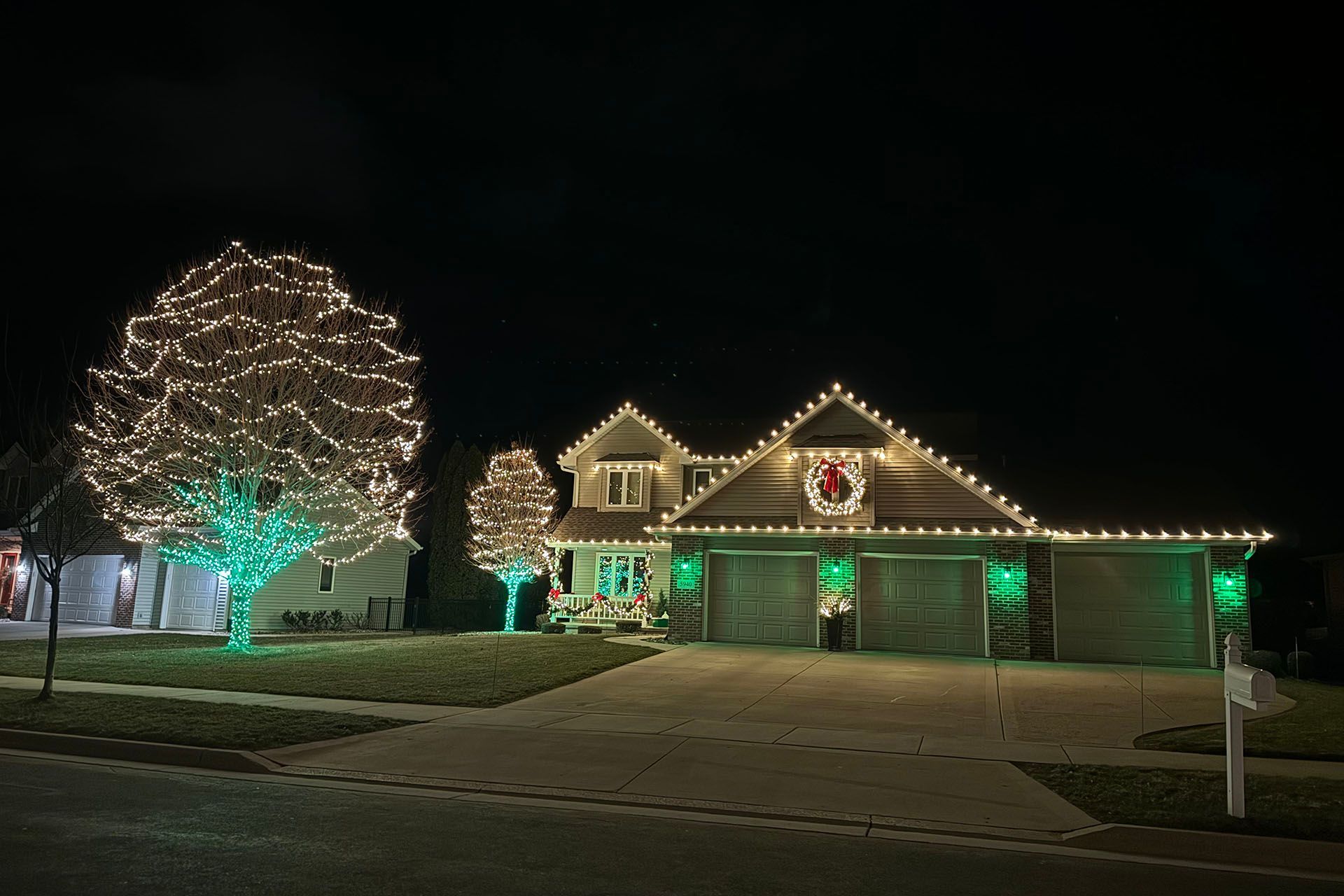 House outlined in white and green holiday lights at night beside a brightly lit tree and lawn