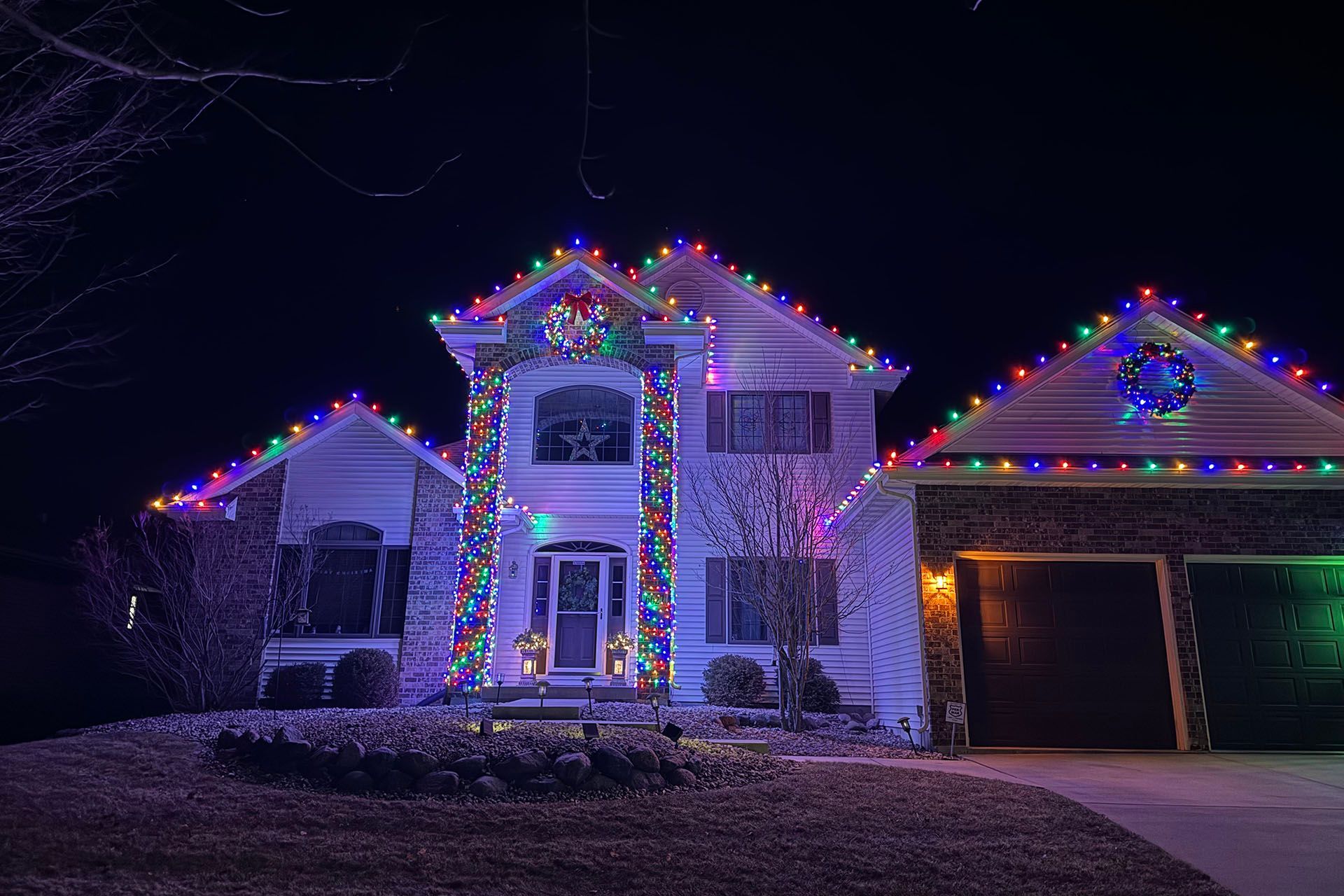 House decorated with colorful Christmas lights at night, with a lit garage and snowy yard