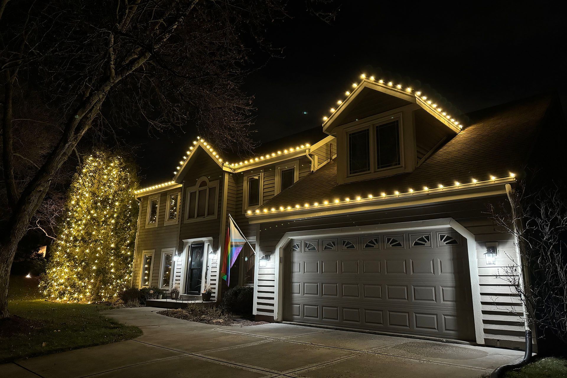 House outlined with warm white holiday lights at night, with a lit tree in the yard and a garage door visible.