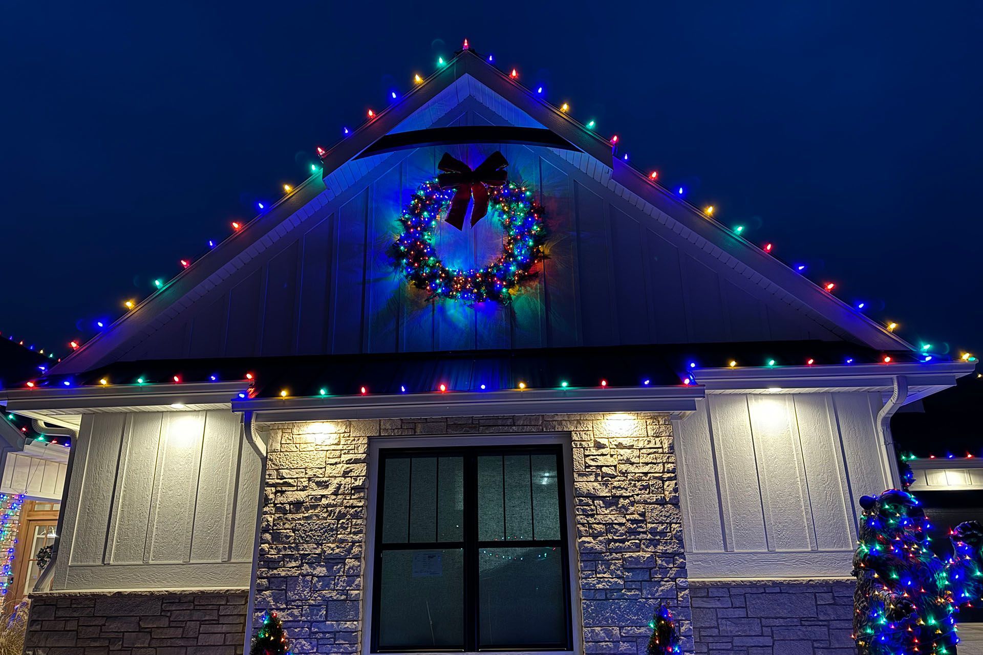 A house roofline and wreath lit with colorful Christmas lights at night