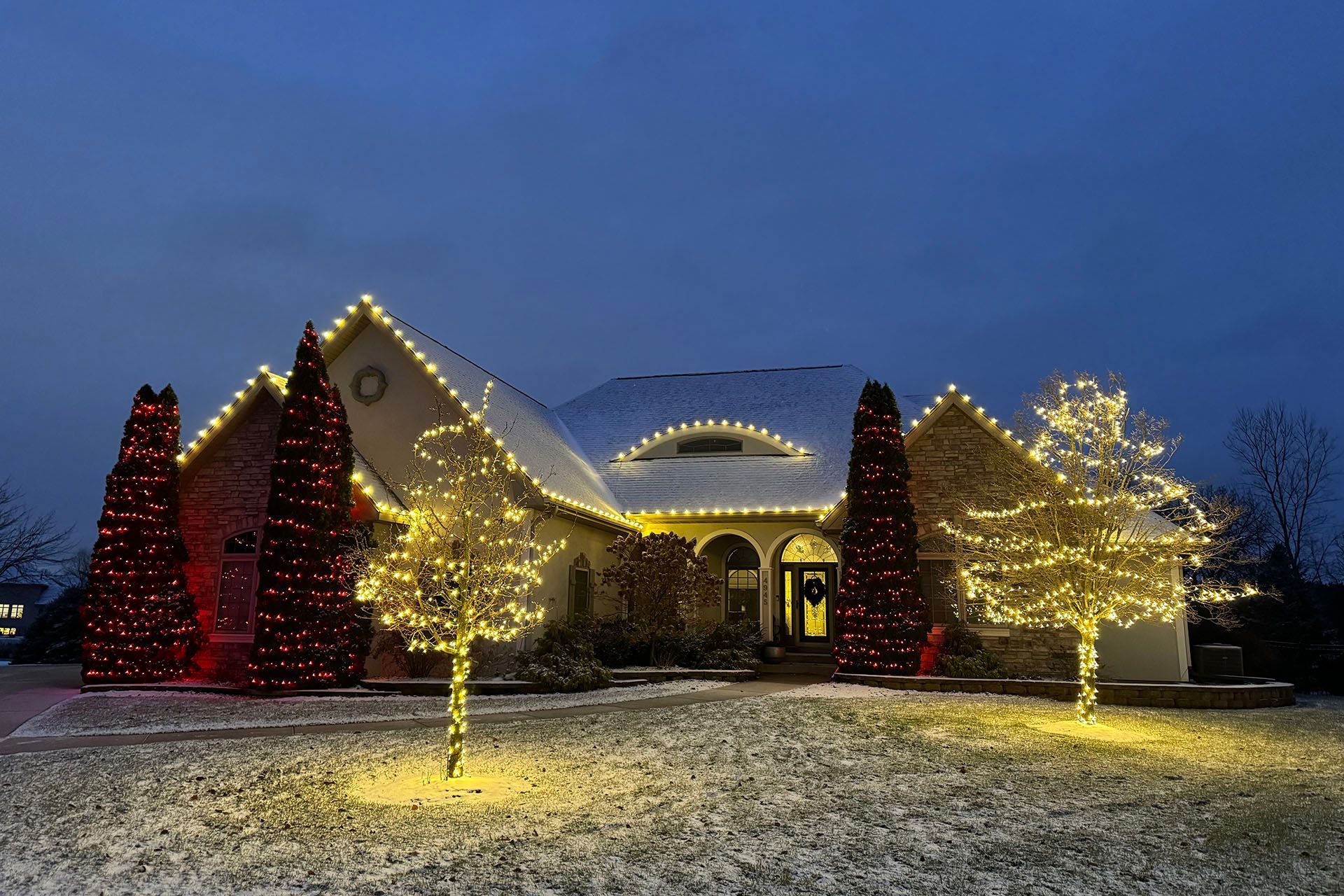 House decorated with holiday lights at dusk, with glowing trees and snow on the ground