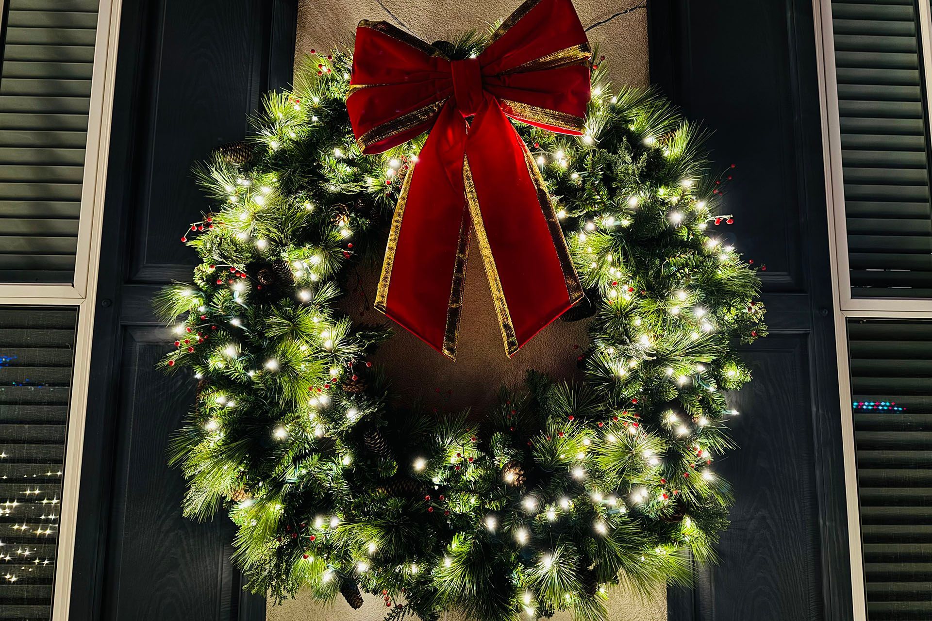 A lit Christmas wreath with a large red bow hanging on a dark window or door.