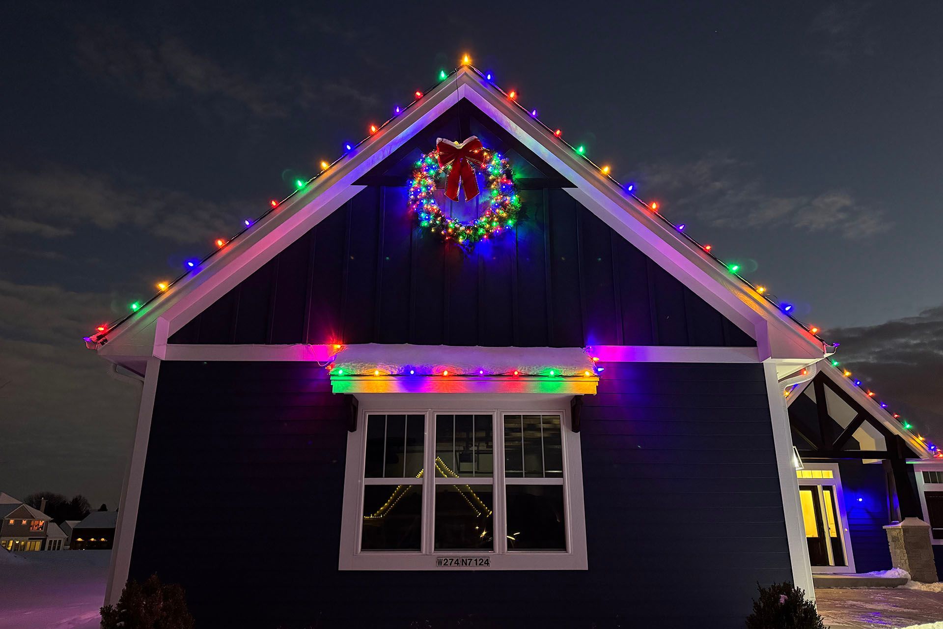 House outlined with colorful Christmas lights at dusk, with a wreath above the window.