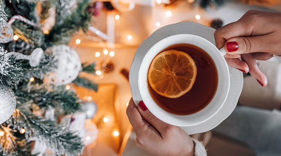 Hands holding a cup of tea with an orange slice beside a lit Christmas tree