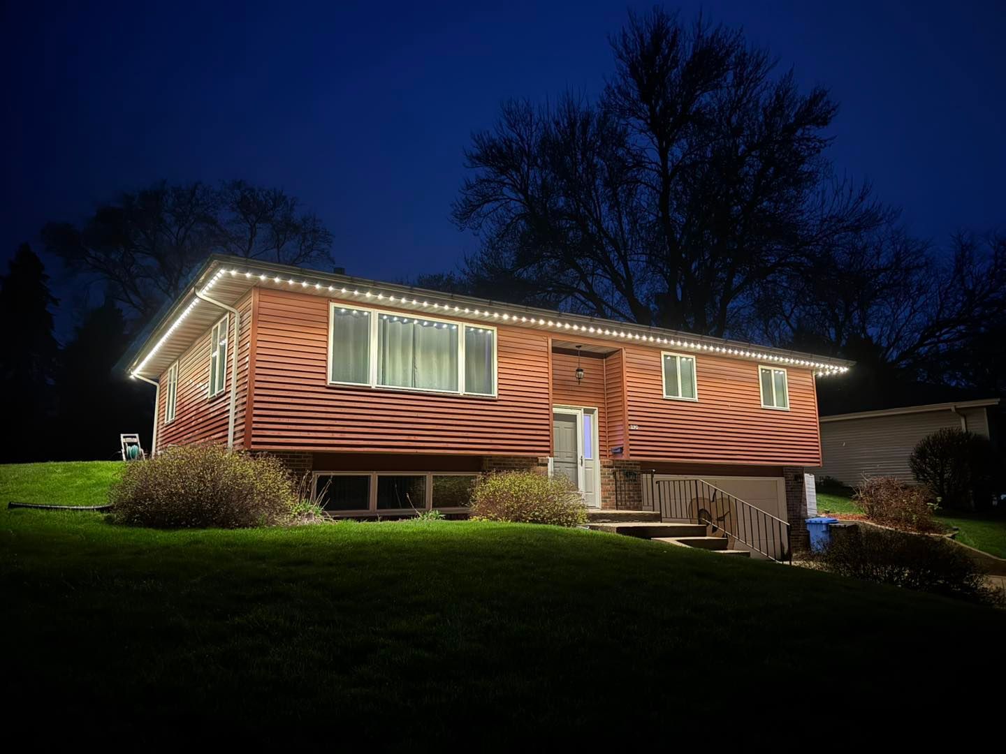 Single-story house at night with red siding, lit windows, and holiday lights along the roofline.