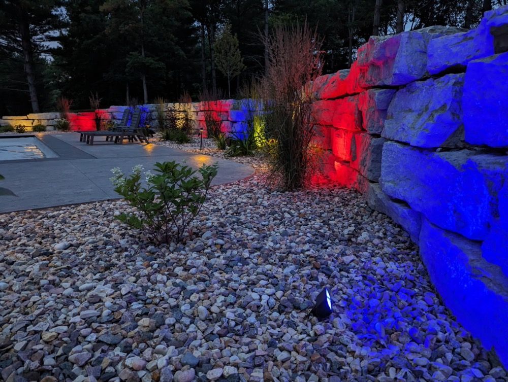 Color-lit stone wall in a landscaped outdoor area at dusk, glowing blue and red beside gravel and shrubs