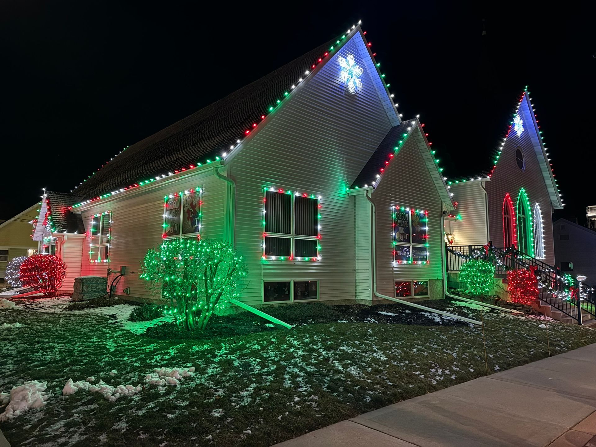 House decorated with colorful Christmas lights at night, with green-lit bushes and snow on the ground