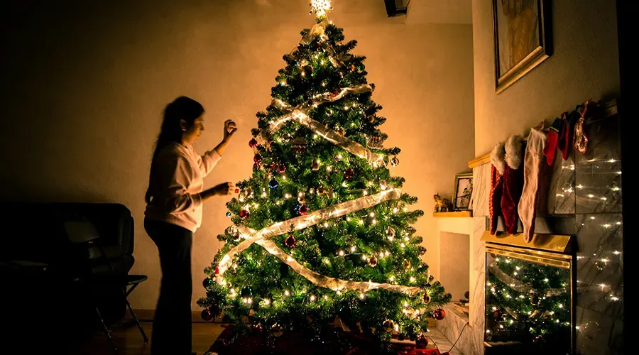 Person decorating a lit Christmas tree in a cozy room with stockings and gifts nearby