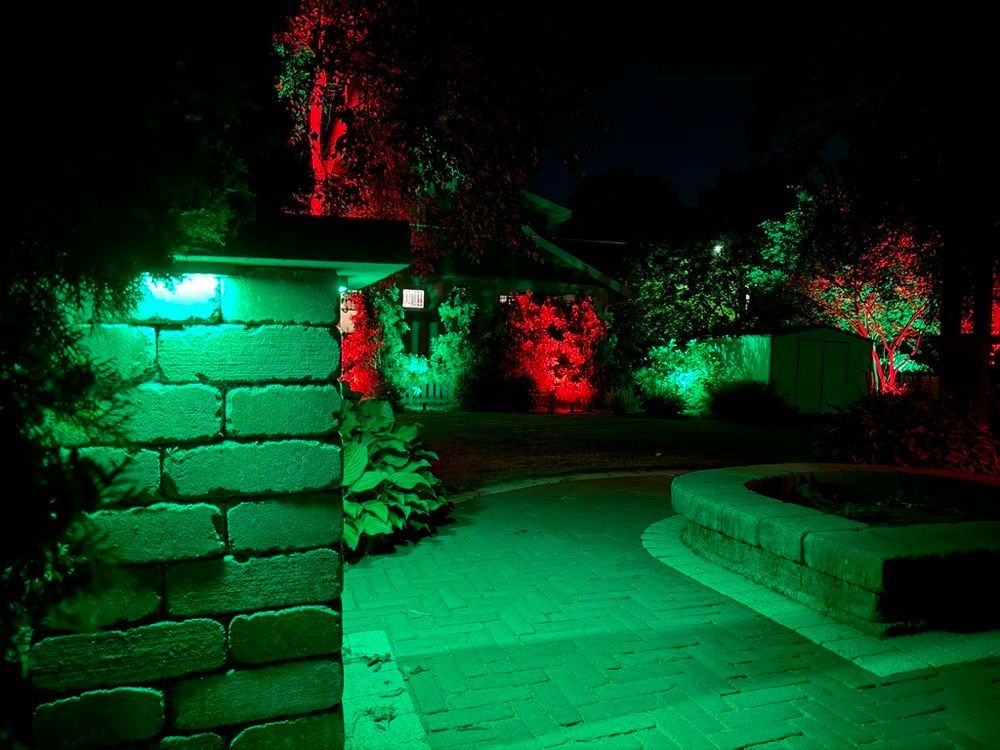 Night scene of a stone pathway and wall lit by green and red lights, with trees and a bench nearby