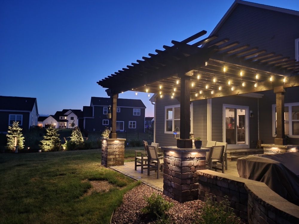 Backyard patio with pergola string lights at dusk, stone columns, outdoor seating, and neighboring houses.