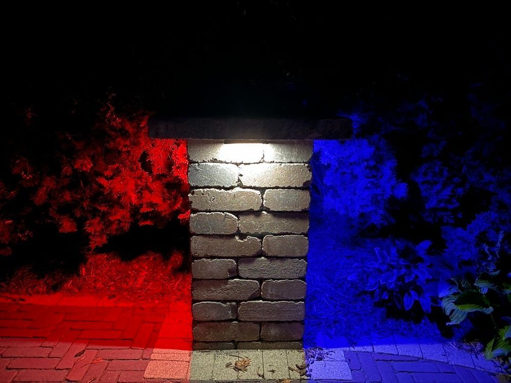 Brick pillar lit white, flanked by red foliage on the left and blue foliage on the right at night
