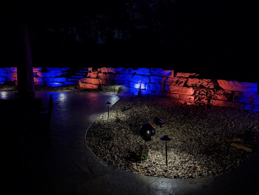 Nighttime outdoor scene with blue and red lit stone wall and a gravel path with a lamp in the foreground