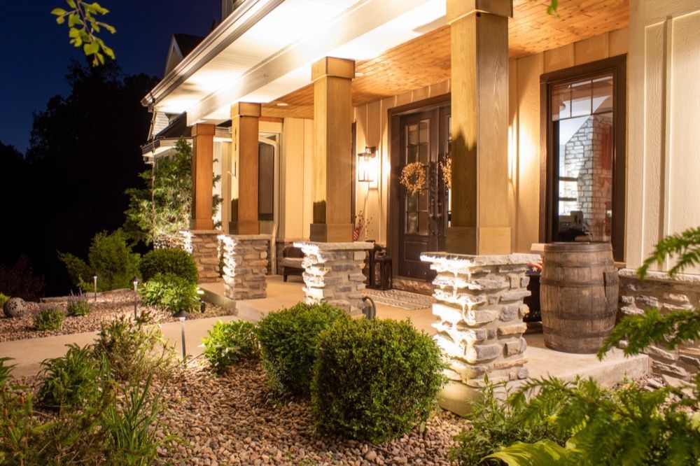 Warmly lit front porch of a house at night with stone columns, shrubs, and landscaped garden.