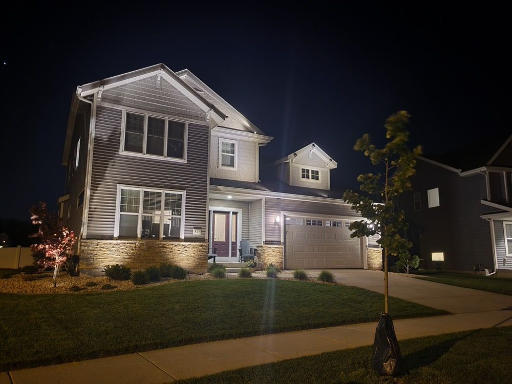 Two-story suburban house at night with porch lights on and a driveway, lawn, and small tree in front.