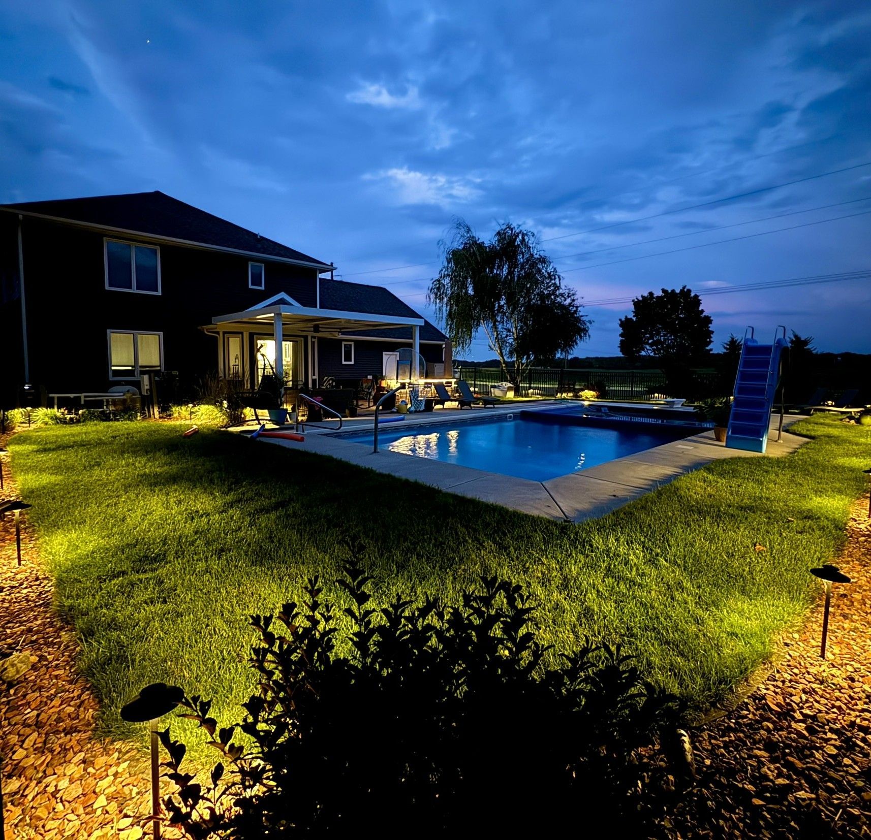 Nighttime backyard with a lit swimming pool beside a house and grassy yard under a blue sky