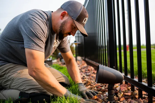 Worker installing garden lights along a black metal fence with rocks and greenery.