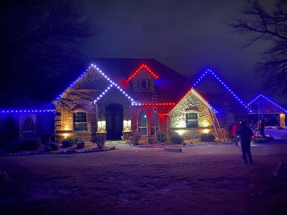 House at night outlined with red, blue, and white holiday lights, with a person standing in the yard