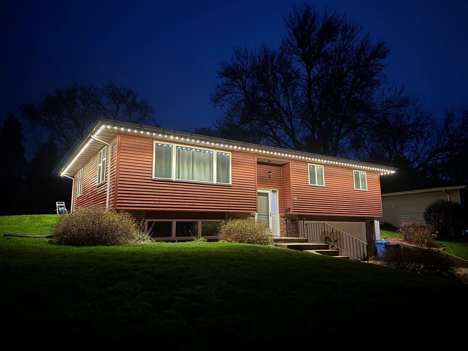 Single-story house with warm exterior lights at dusk, on a grassy lawn beneath dark trees.