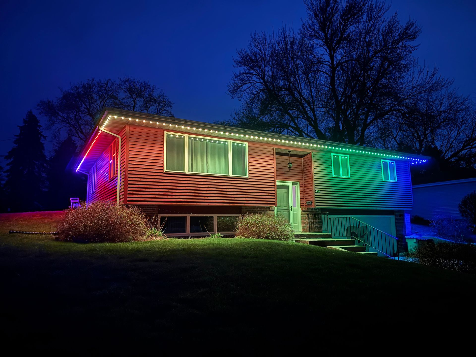 House lit with colorful rainbow lights at night, surrounded by dark trees and a grassy lawn.