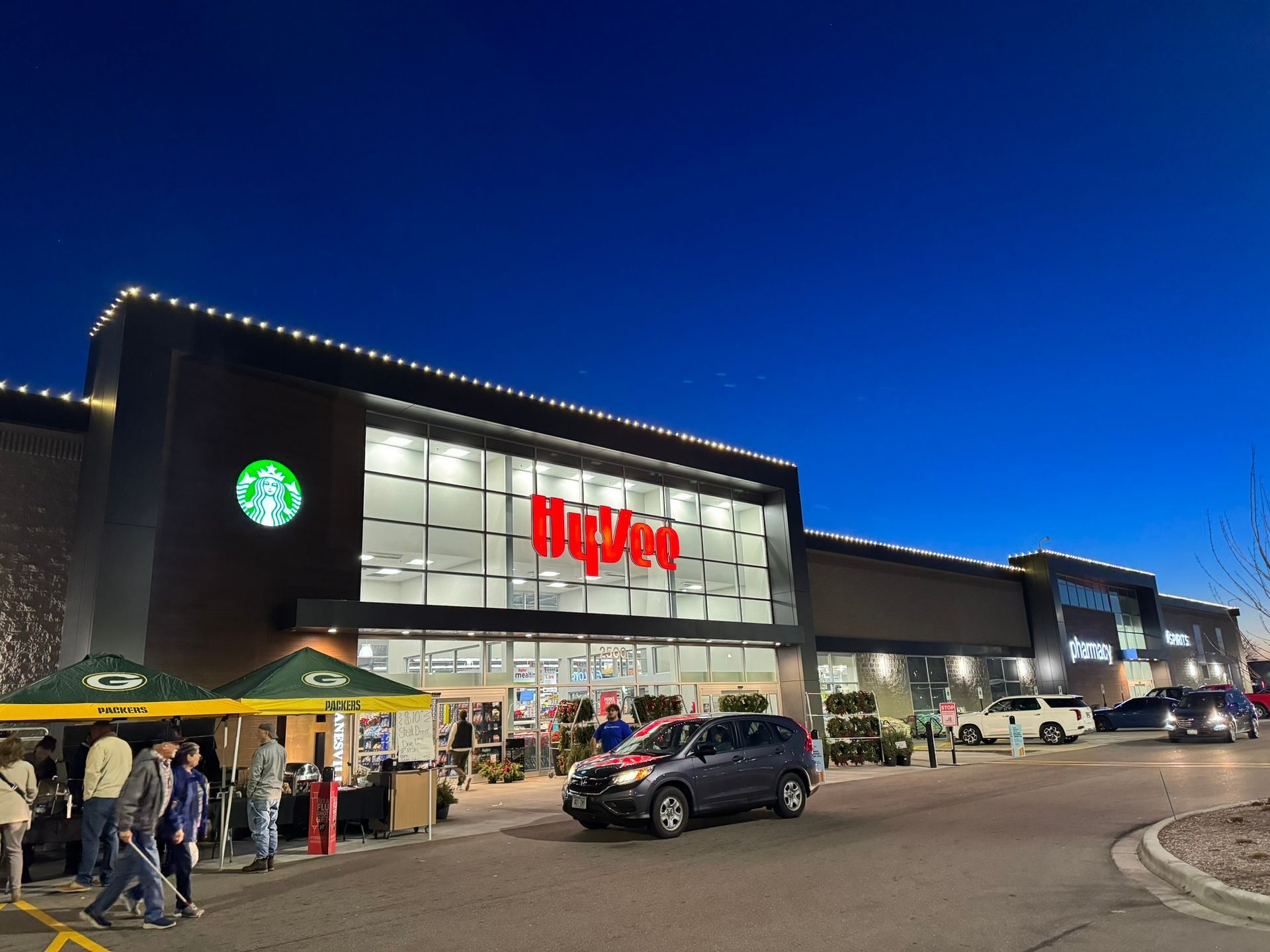 Shopping center entrance at dusk with a Starbucks sign, red store logo, parked cars, and people outside.