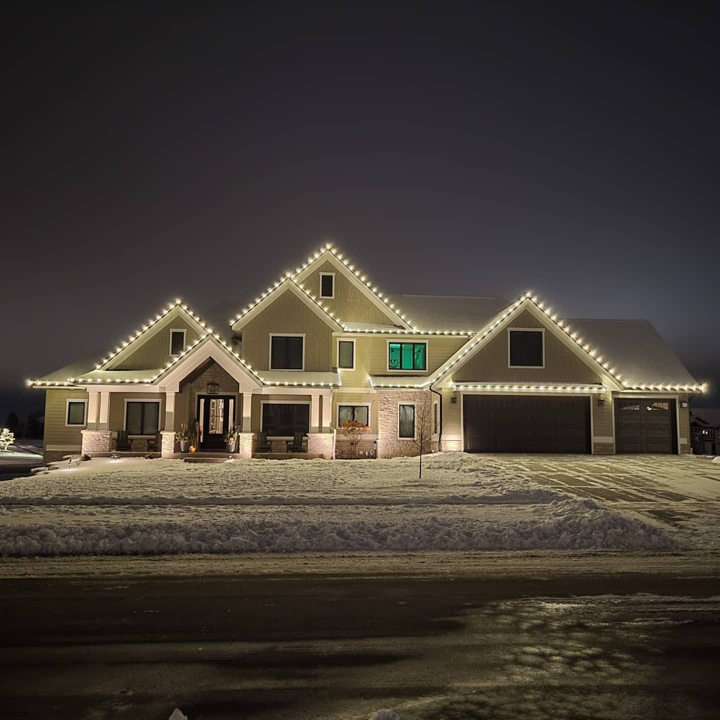 Snow-covered house at night with warm white roof lights and a glowing green window