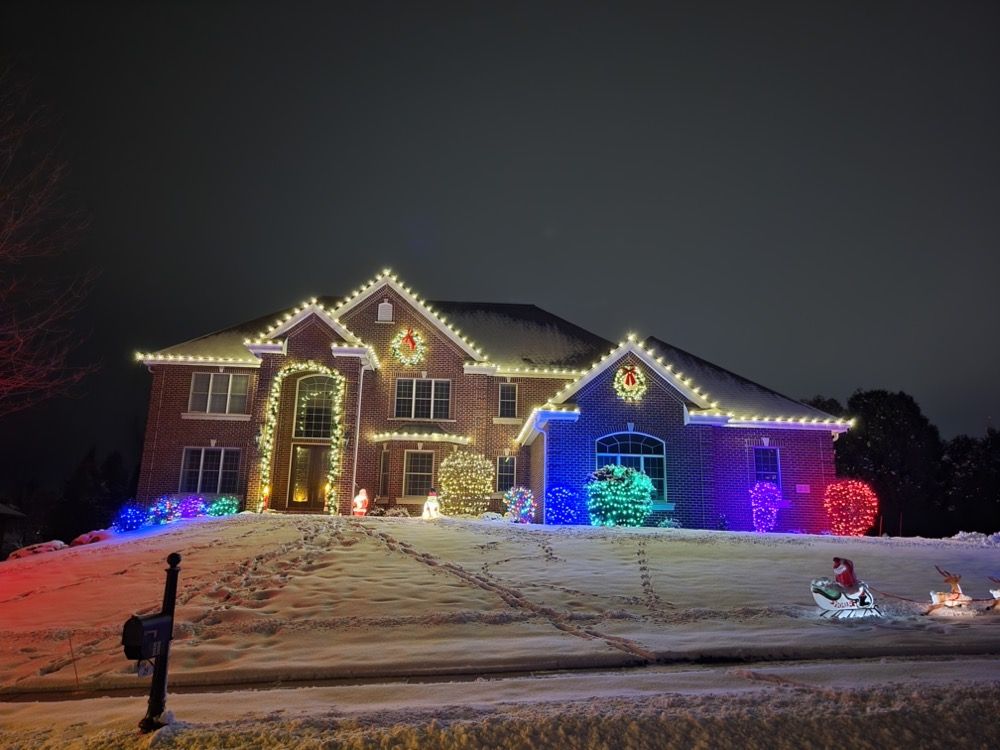Brick house at night decorated with colorful Christmas lights on a snowy front lawn.