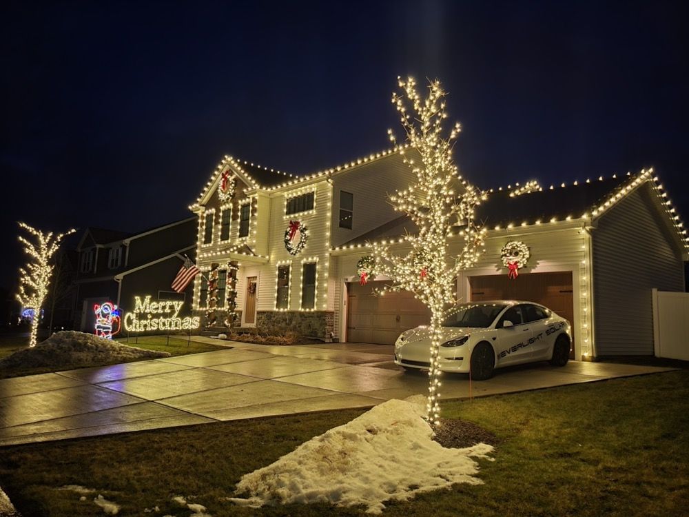 Christmas-lit house with a decorated tree, “Merry Christmas” sign, and a white car in the driveway at night