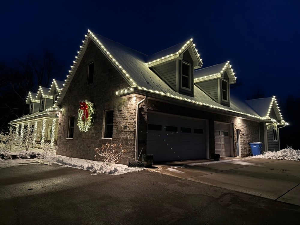 Snowy brick house at night, outlined with warm white holiday lights and a wreath on the garage.