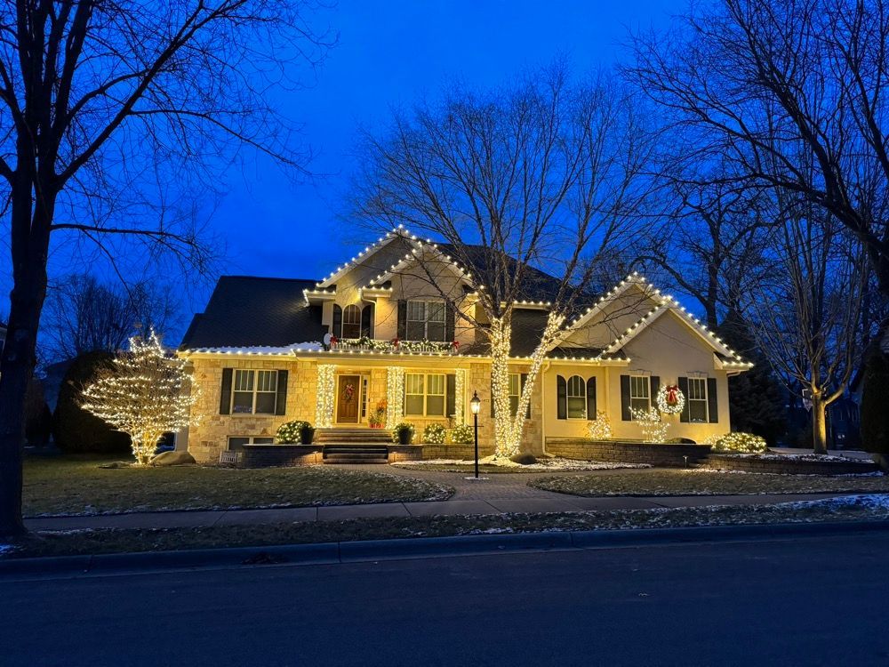Lit house with holiday lights at dusk, surrounded by bare trees and a snowy yard.