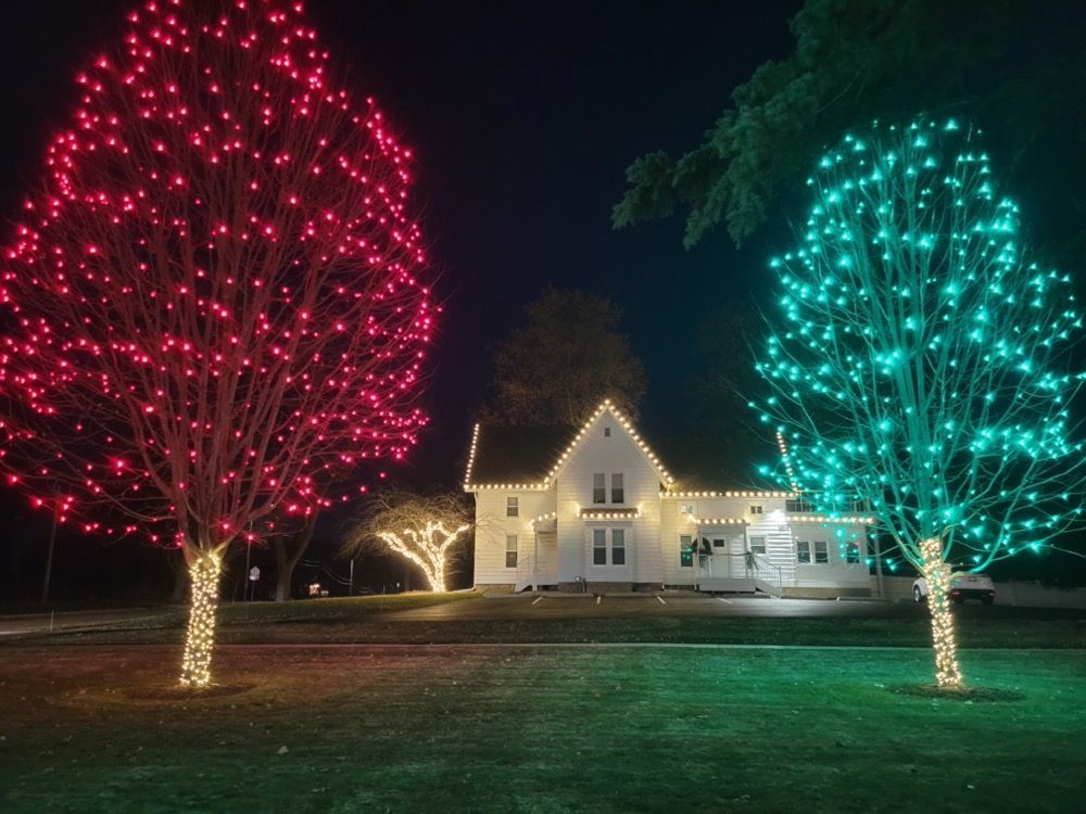 House lit with red and teal holiday lights between two illuminated trees at night
