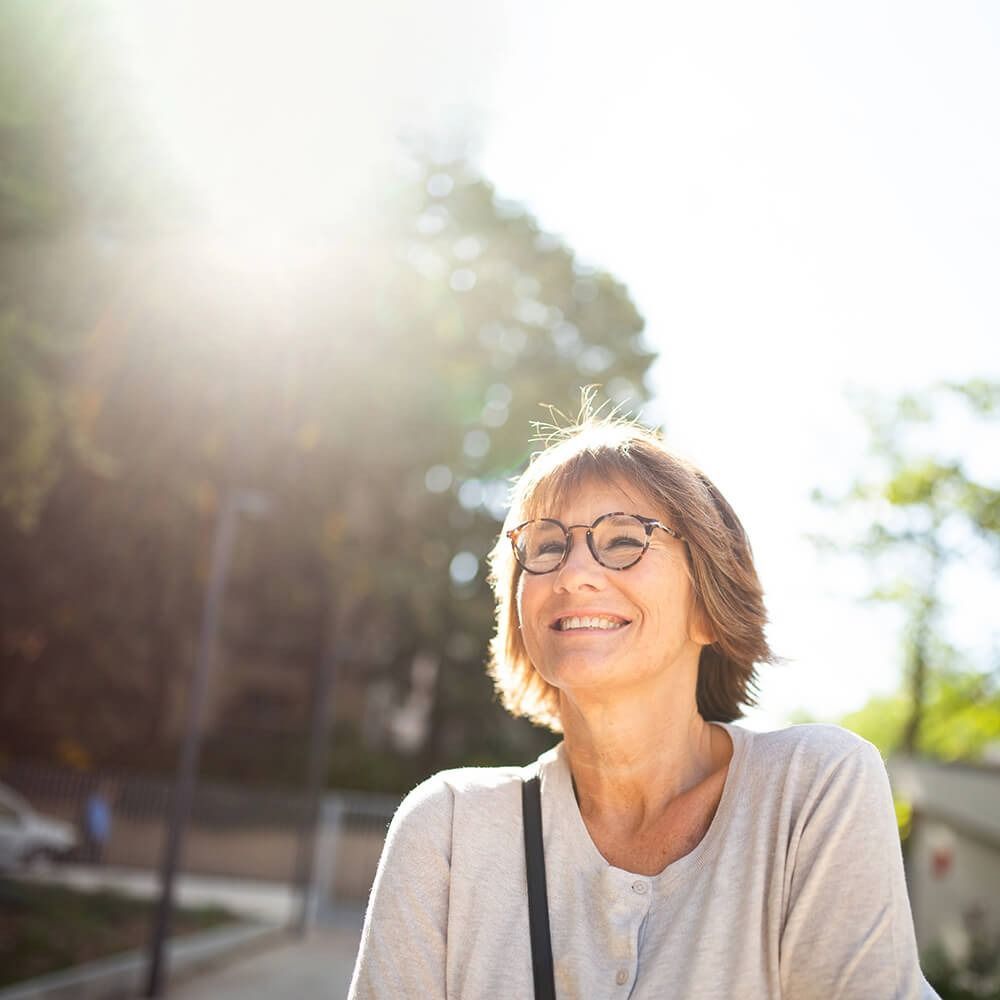 Smiling person with glasses outdoors in bright sunlight, with trees and a building in the background