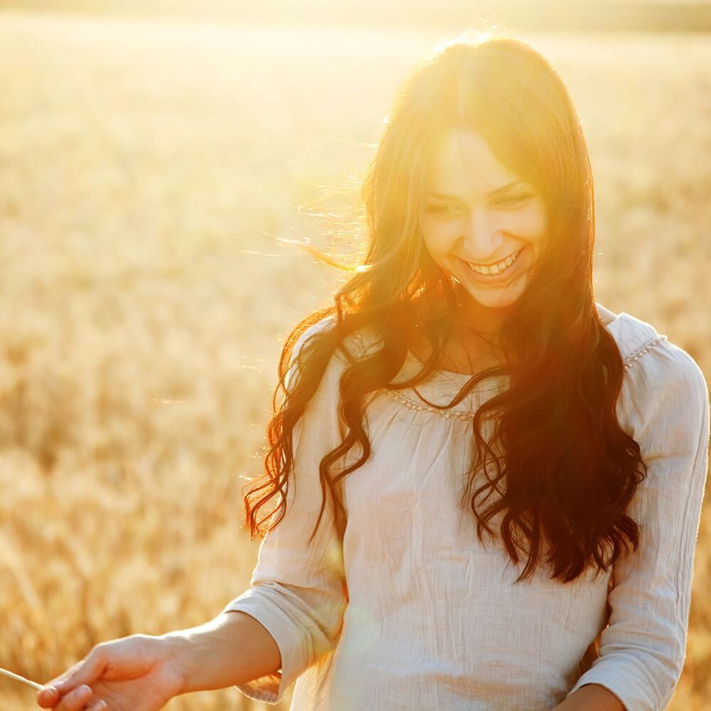 Smiling person in a sunlit field, with long hair and a white top, holding out an open hand