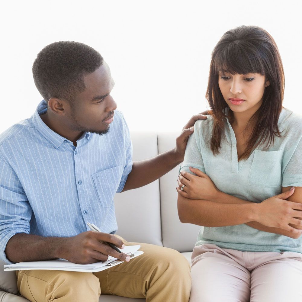 Two people sit on a couch; one comforts the other with a hand on her shoulder.