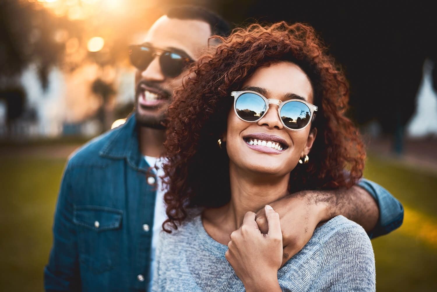 Two smiling people wearing sunglasses outdoors at sunset, one standing behind the other.