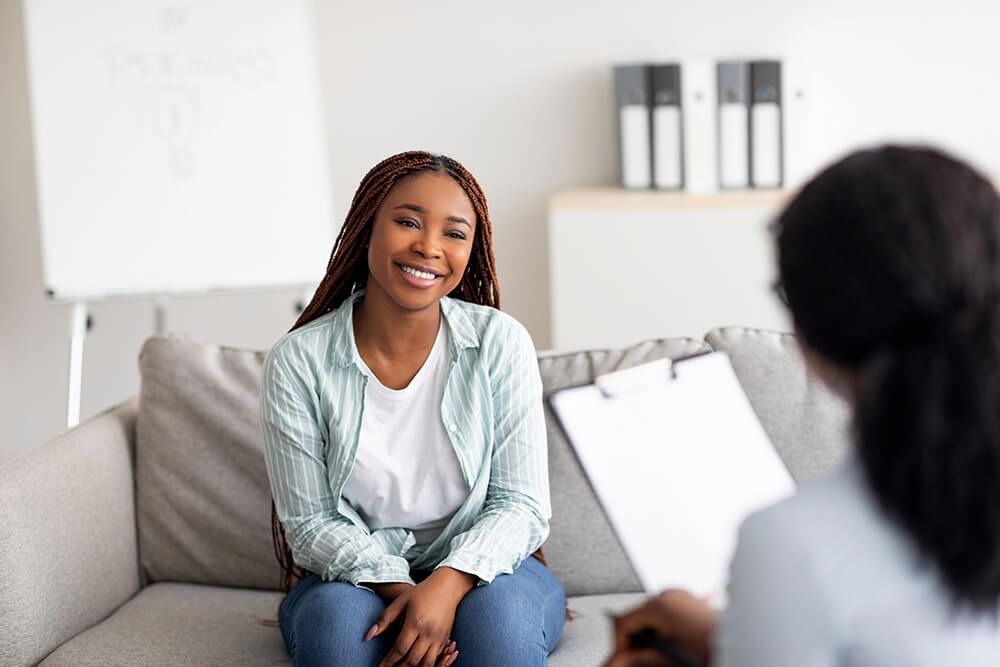 Two people talking on a light gray couch in a bright living room, one smiling and leaning forward