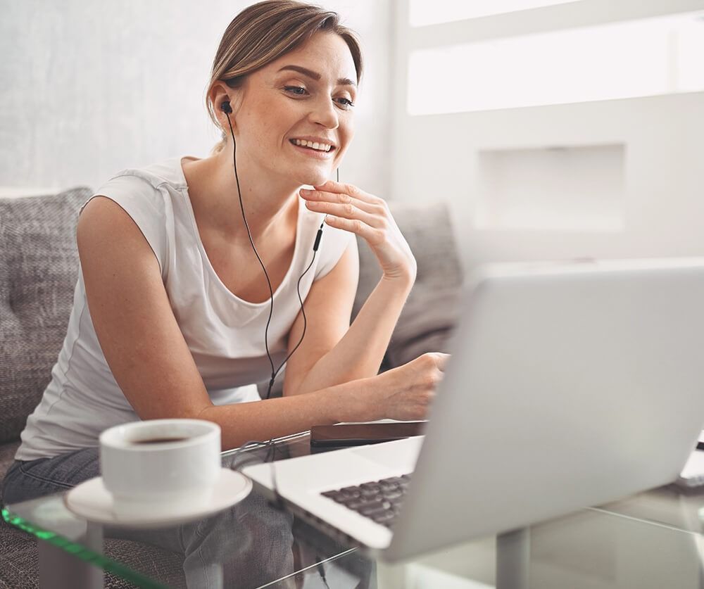 Woman smiling on a couch, using a laptop and wearing earbuds beside a cup of coffee.