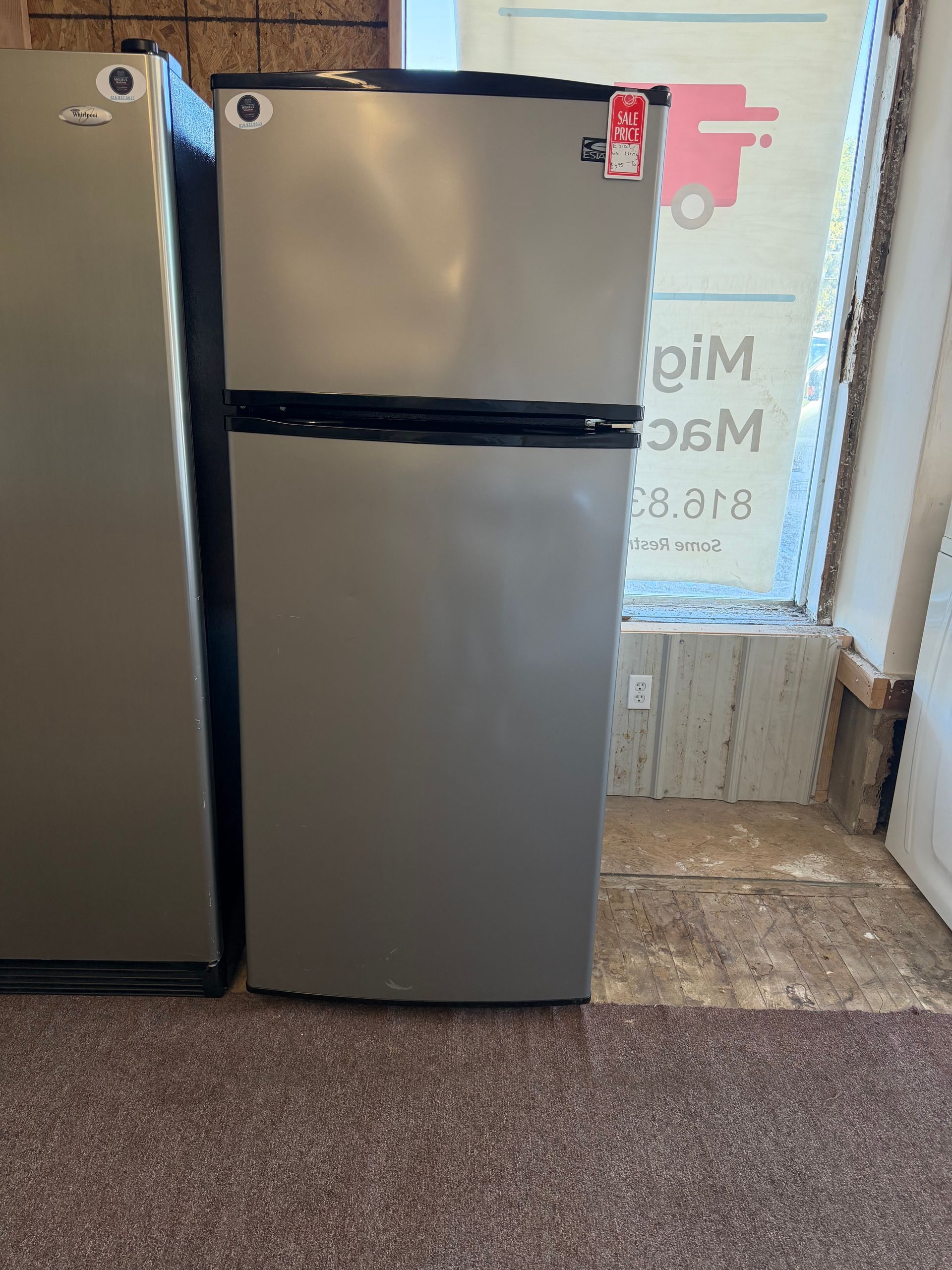 Stainless steel top-freezer refrigerator beside another fridge in a carpeted indoor space