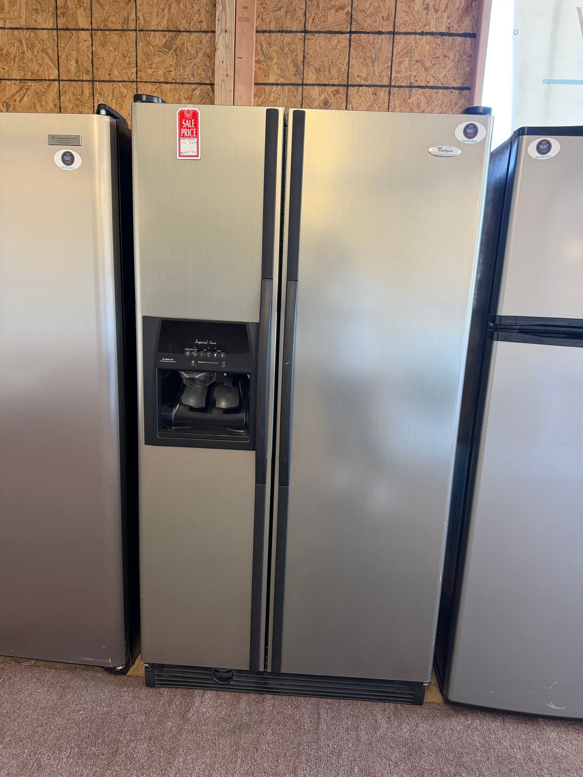 Three stainless steel refrigerators, one with a water dispenser, against a cork wall.