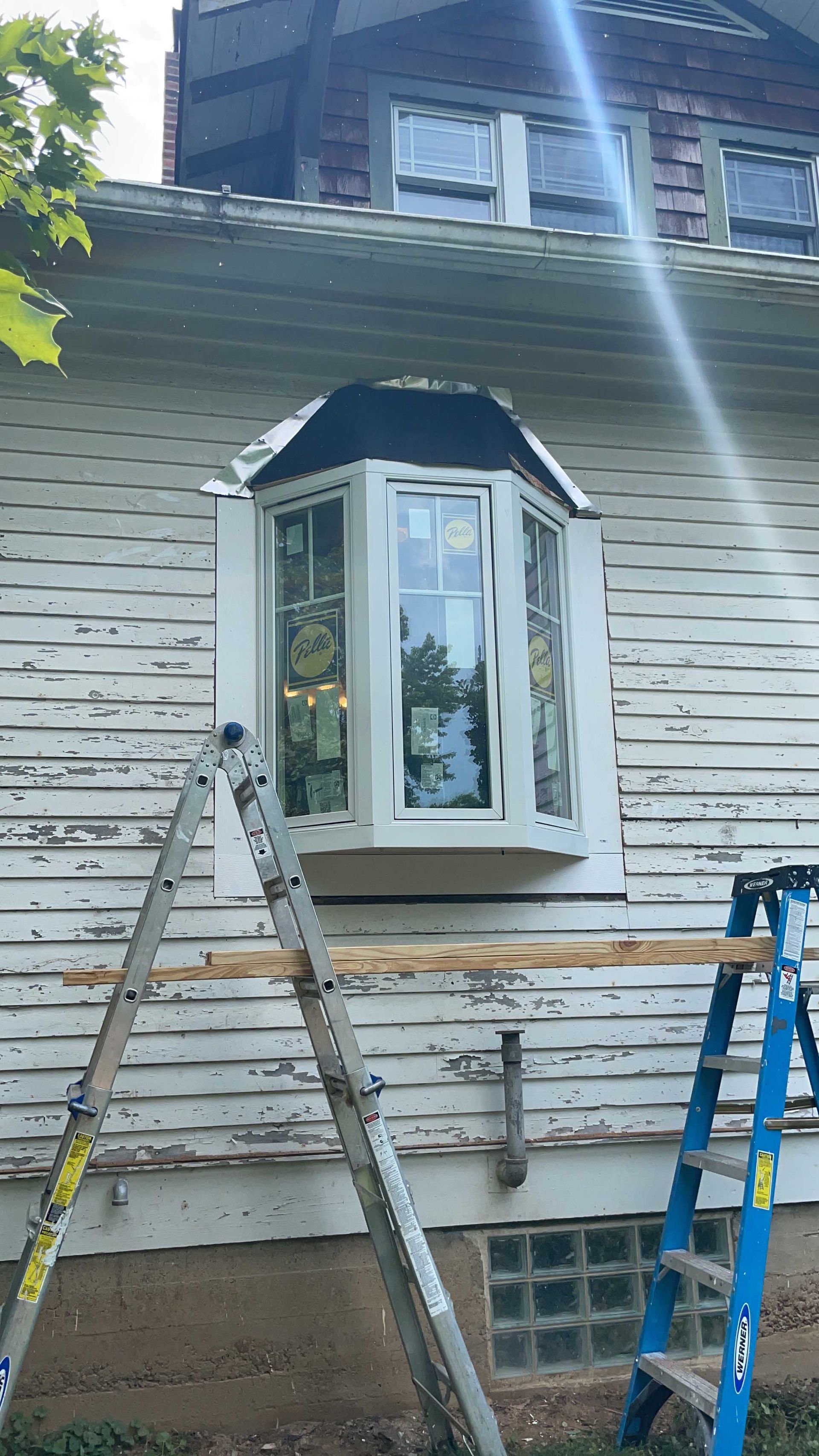 Bay window on a house exterior with ladders propped against the weathered siding.