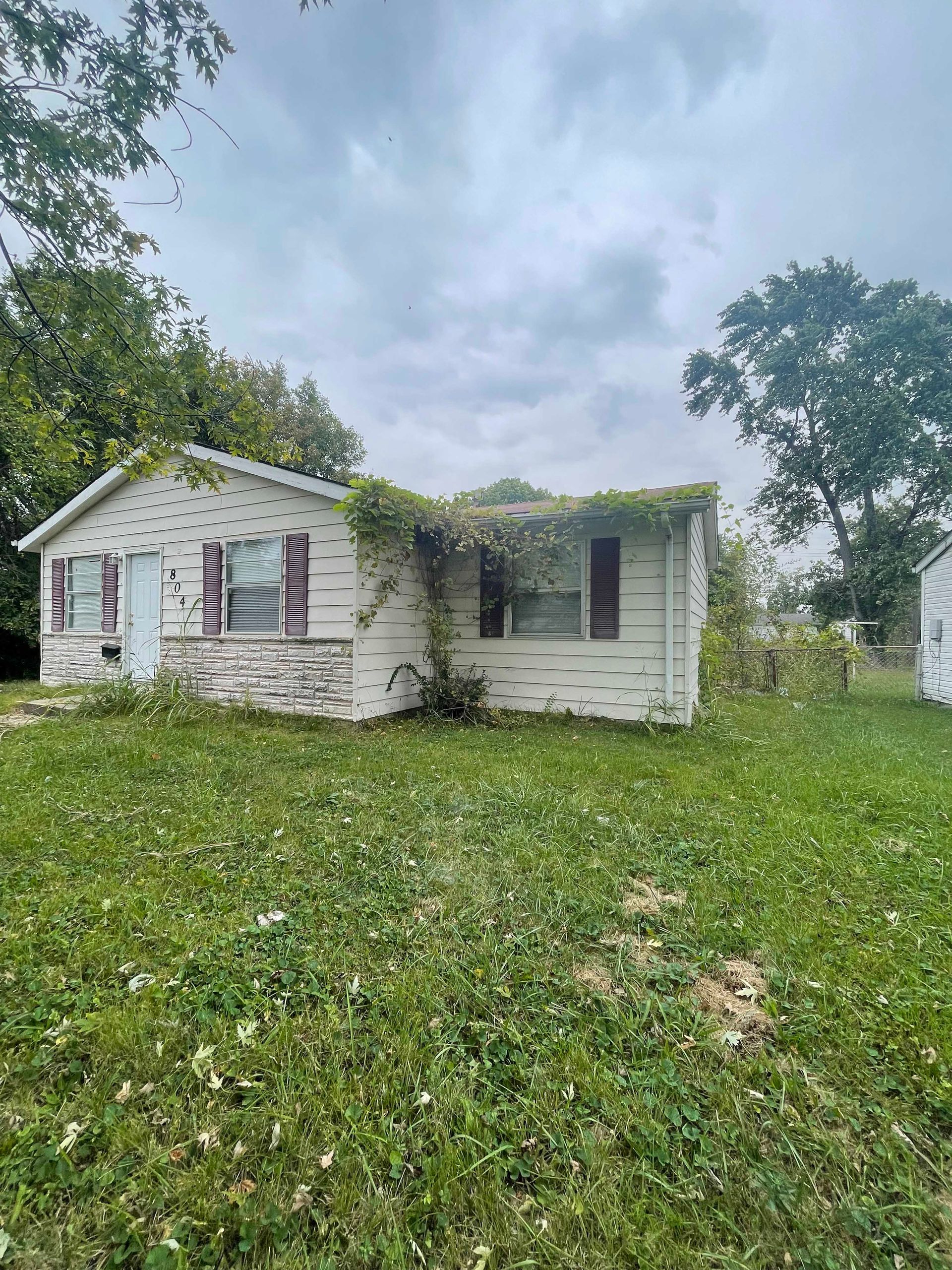 Dilapidated white house with peeling paint and overgrown vines on a cloudy day. Green grass and trees.