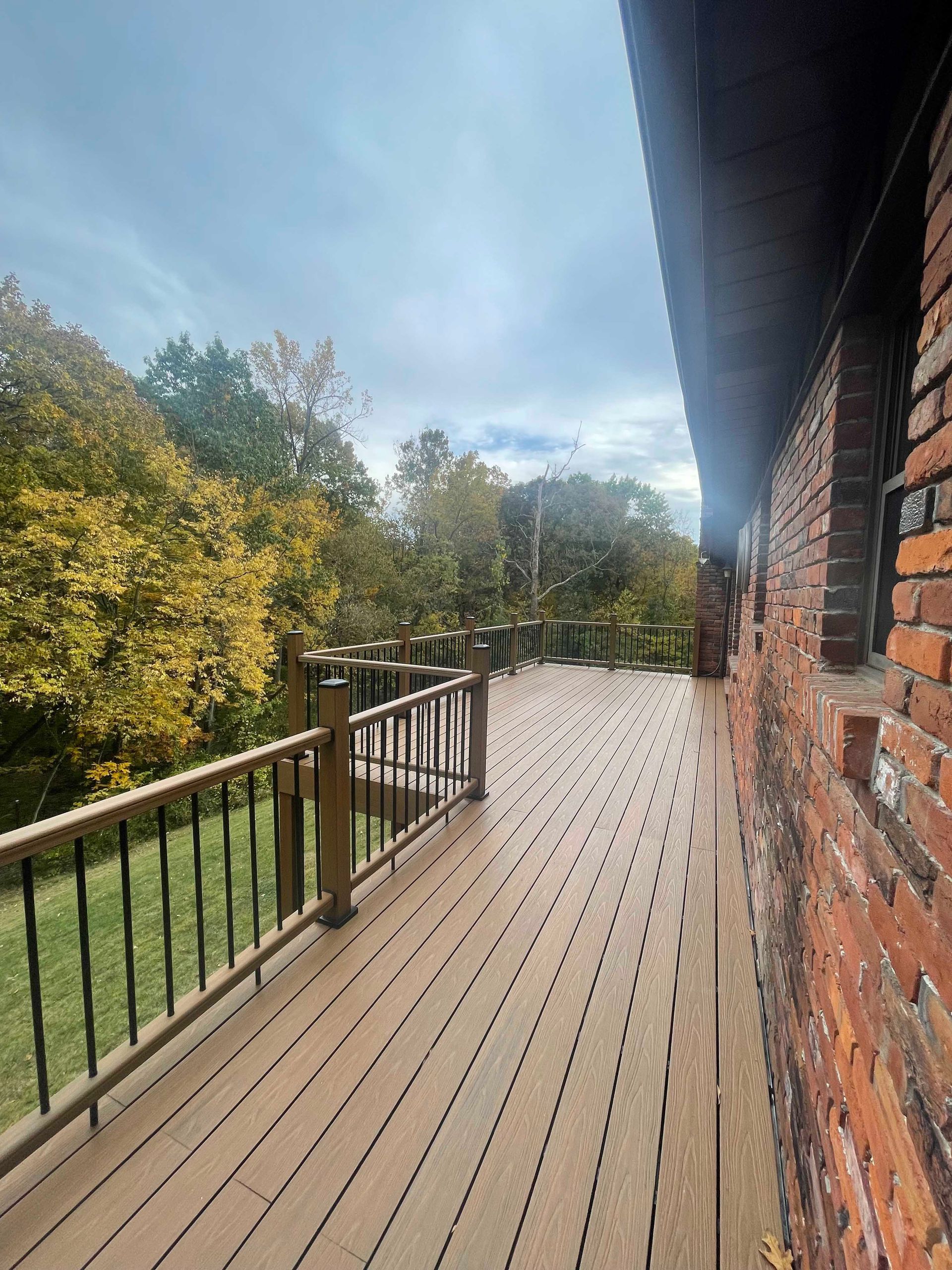 Wooden deck next to a brick building overlooking a yard and trees with yellowing leaves.