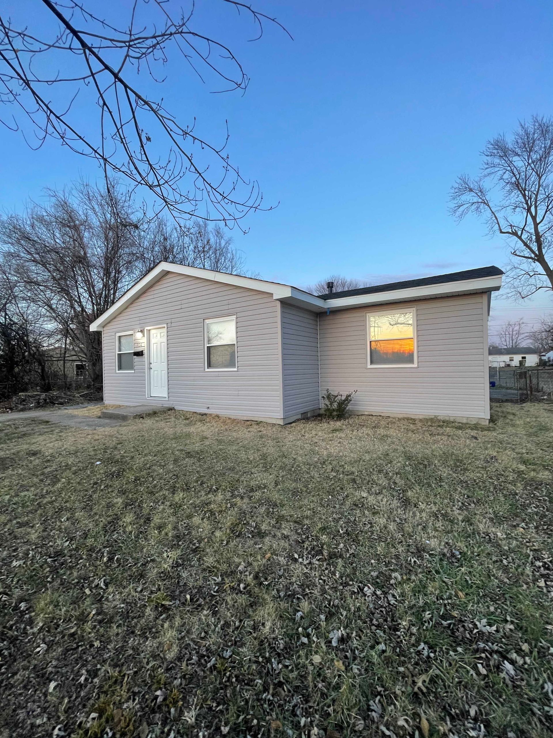 Small gray house with white trim, set on grassy lot under blue sky, bare trees.