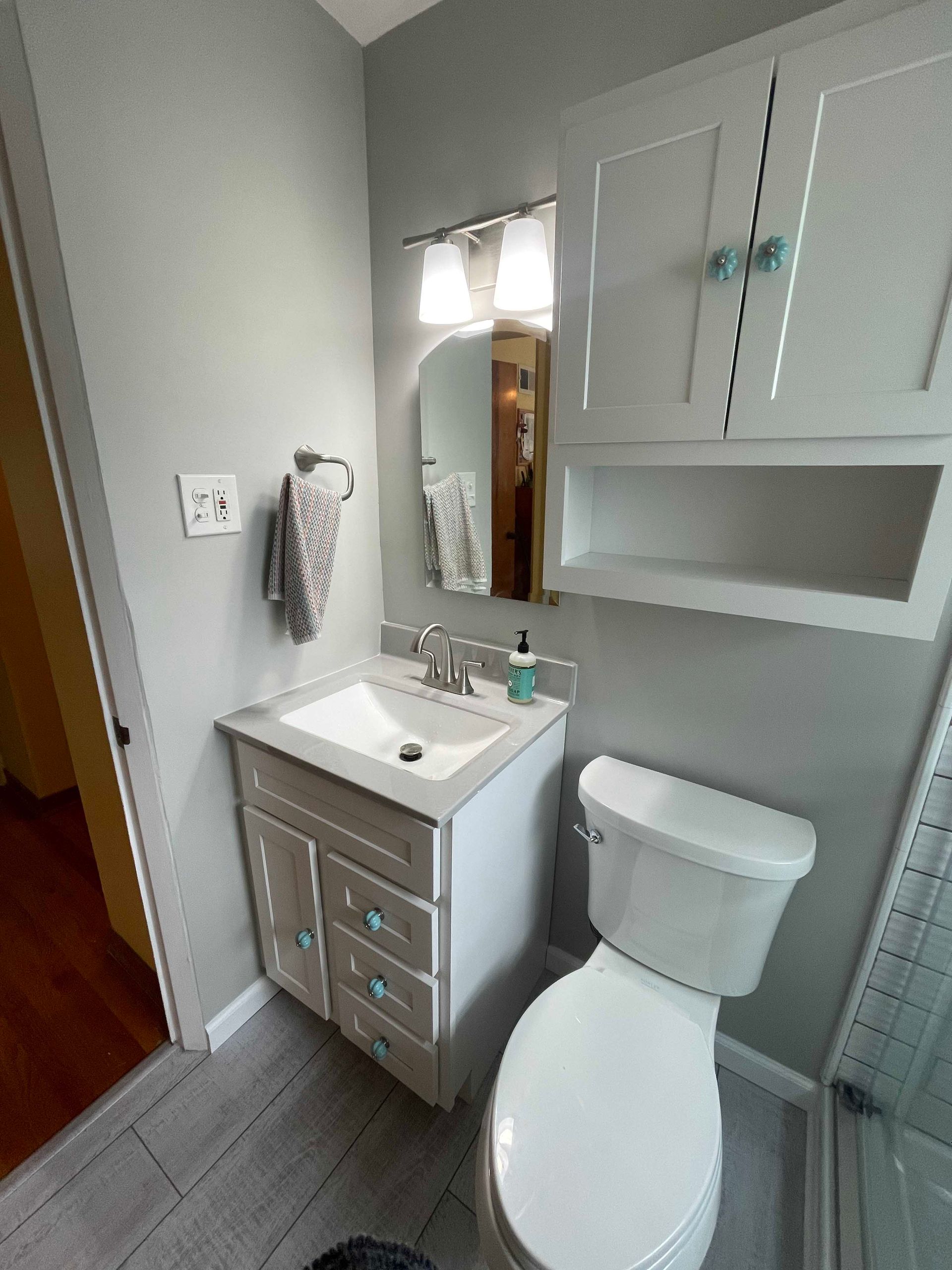 A small bathroom with gray walls and a white vanity, toilet, and cabinet with teal knobs.