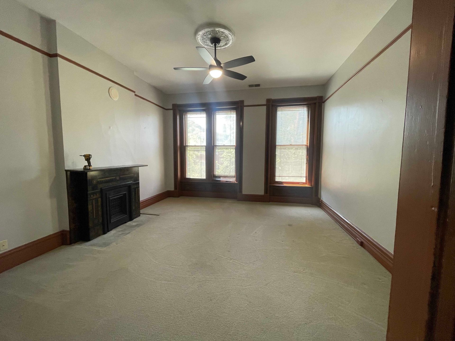 Empty room with fireplace, windows, and ceiling fan. Gray walls, brown trim, and carpeted floor.