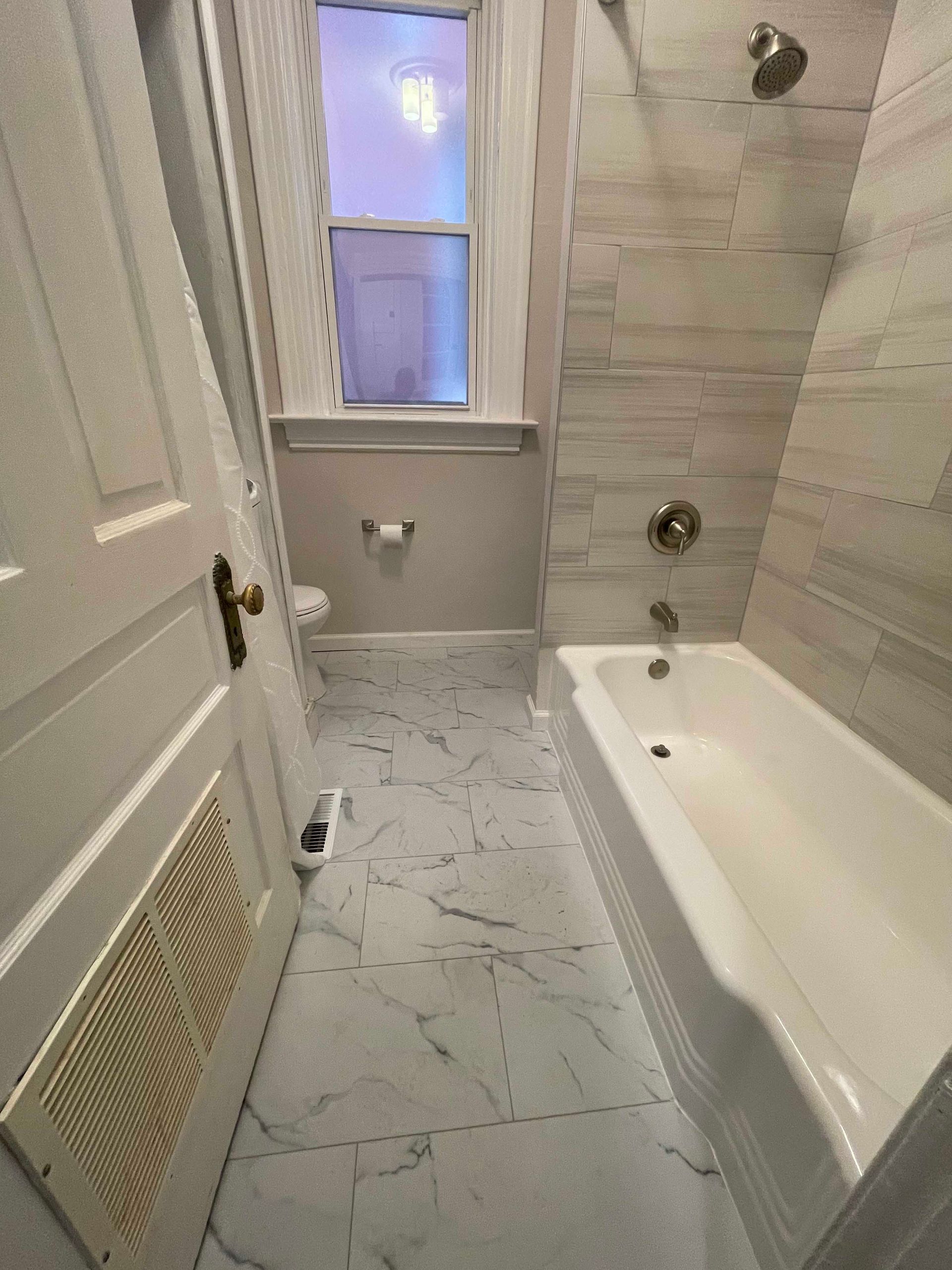 Bathroom with white tile flooring and wall-mounted showerhead above a bathtub. Light enters from a window.