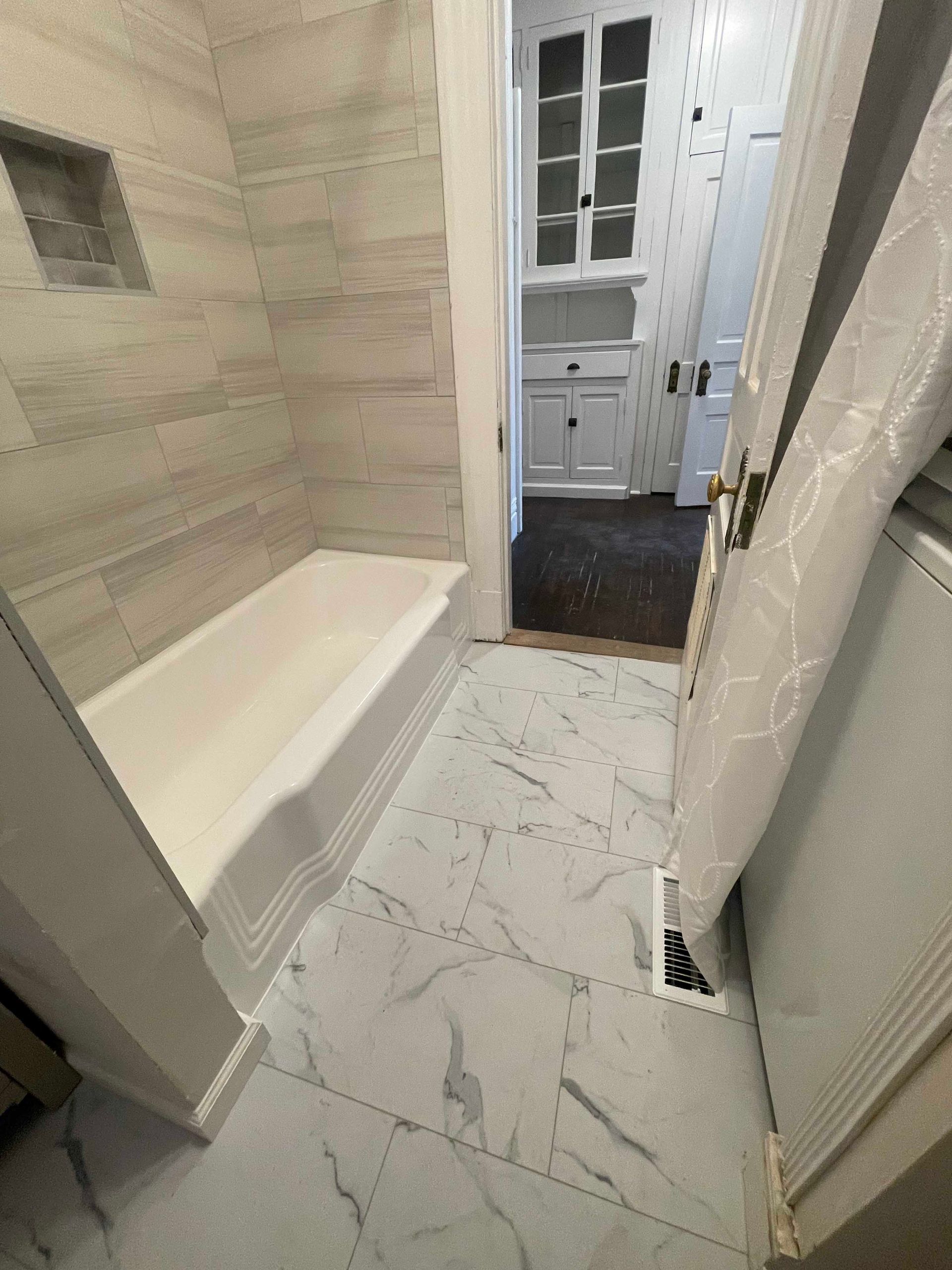 Bathroom with marble floor, white tub, and light-colored tile walls. Door leads to another room.