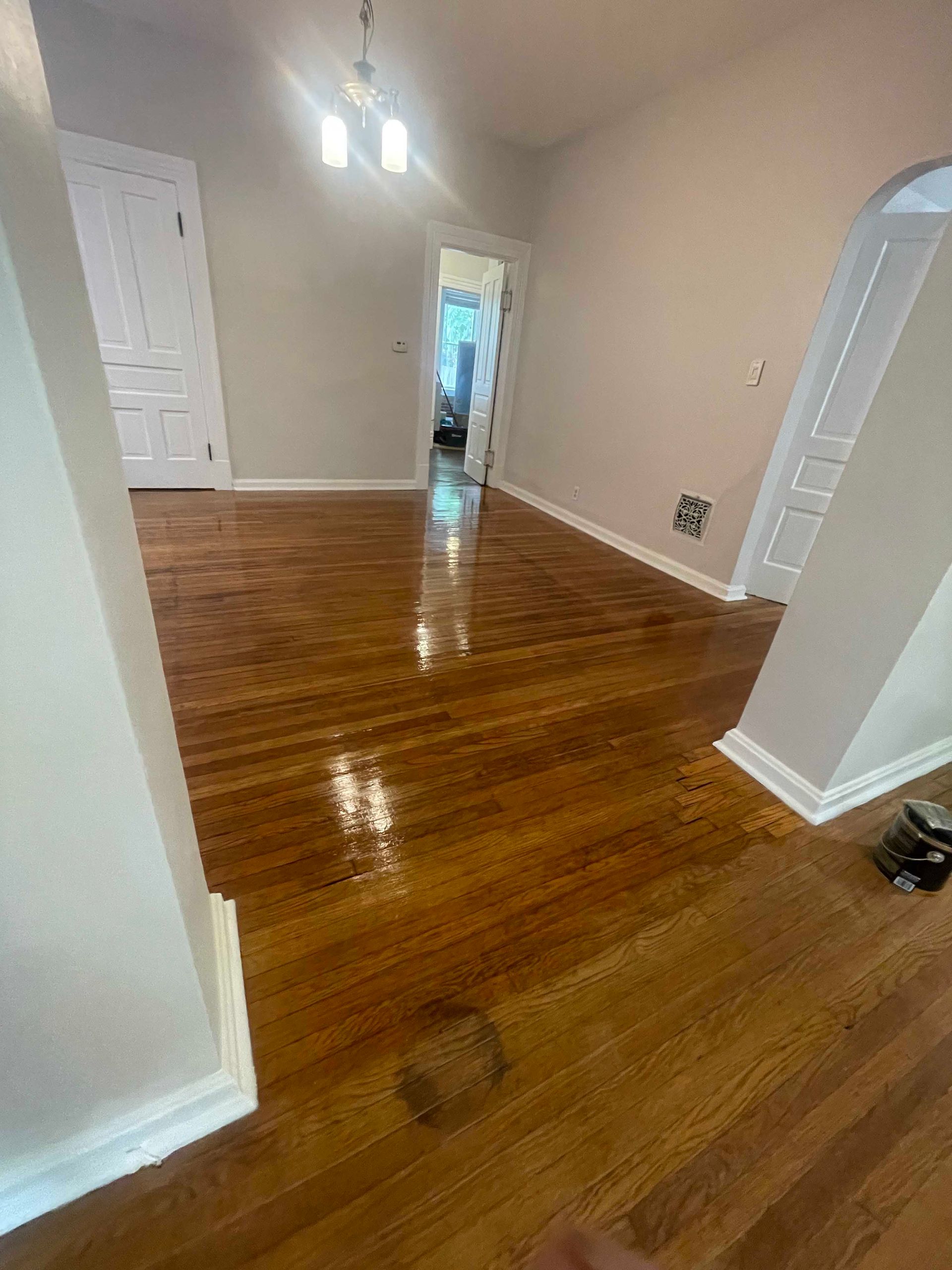 Wooden floors in a room, with a glimpse into a hallway. Light walls and a chandelier are visible.
