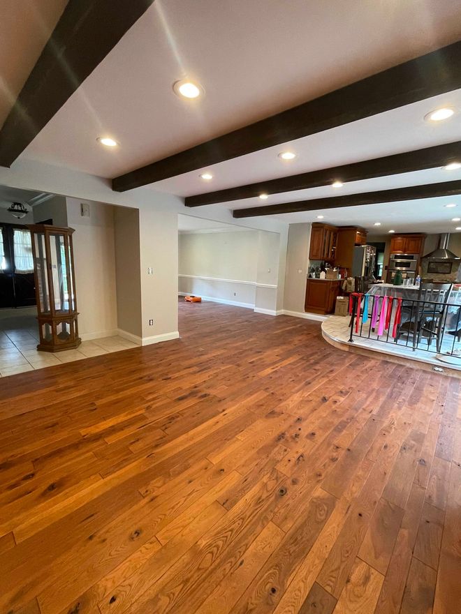 Spacious room with wood floor, dark ceiling beams, and recessed lighting. Kitchen visible in the background.